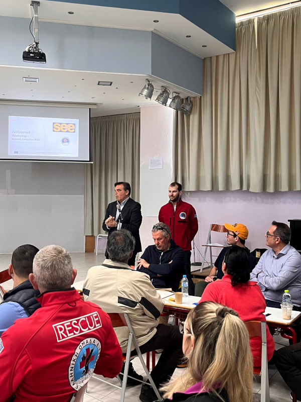 In the foreground, there are a number of people sitting at tables, looking to the other end of the room where there are two speakers standing up and apparently talking. Behind them, there is a projection identifying the meeting as part of the SEE project.
