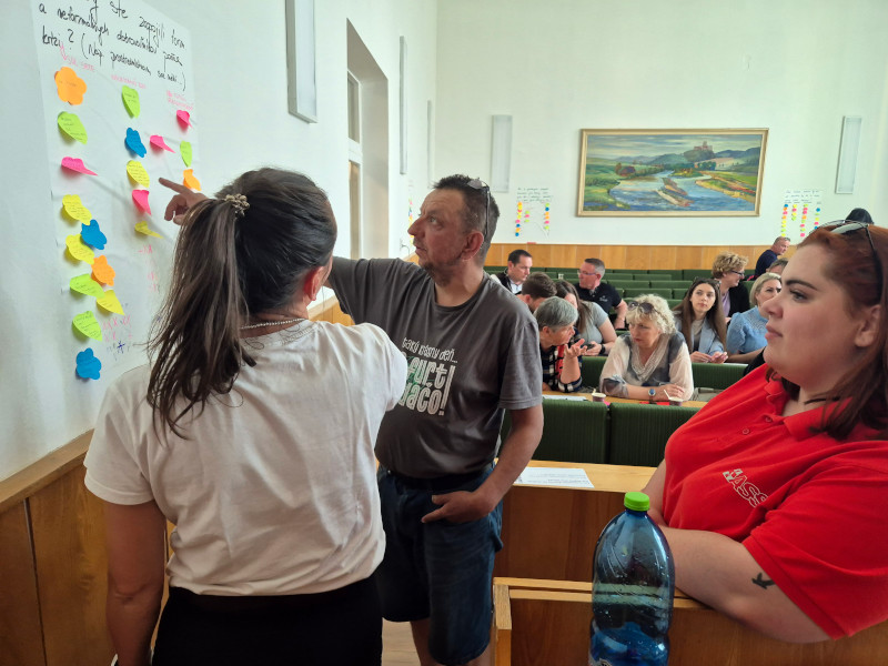 Three people, two women and one man,  in the foreground are discussing and working on a Flipchart page pinned to the wall to their left. In the background, there are several rows of plenary-style seating with people in them, some of them can be seen talking with each other.