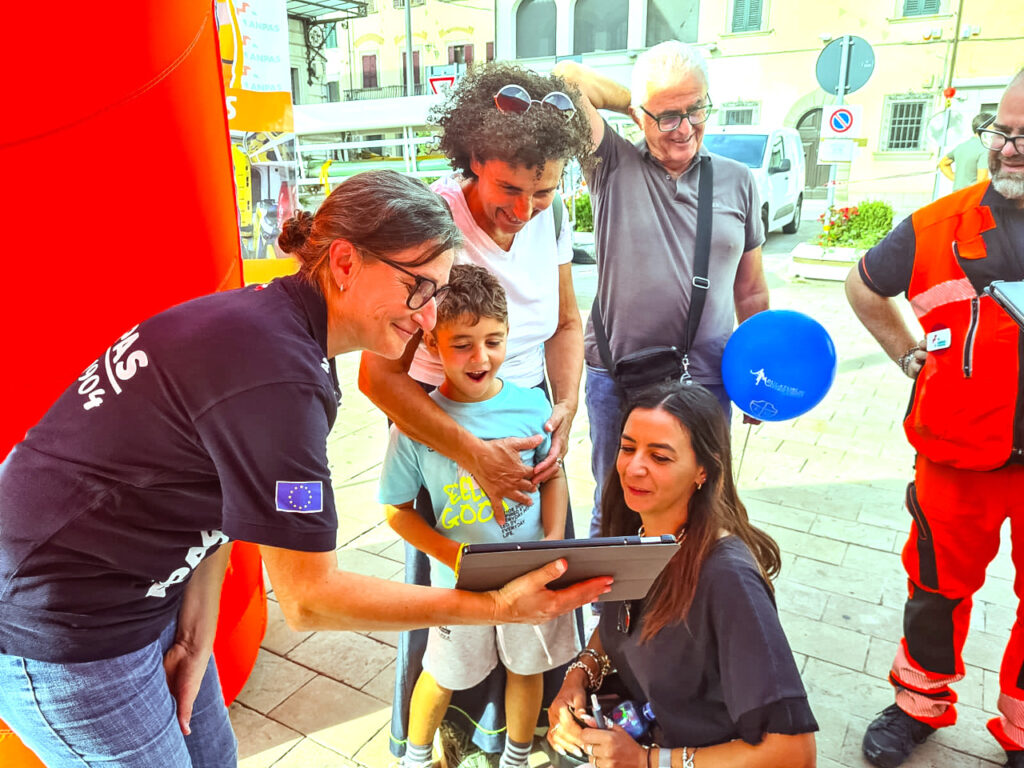 Photograph of an ANPAS volunteer explaining something to a group of people, a child, two women and a man, by showing it on a tablet computer. On the right, there's anther ANPAS volunteer looking on.
