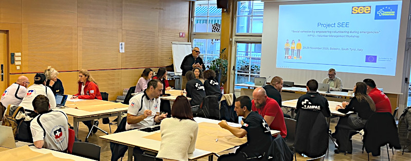 Panoramic view of one half of a large-looking meeting room. People are sitting around several smaller tables and appear to be in discussion. Some of the participants in the picture can be identified as belonging to different aid organisations from their clothing. There is a projection screen in the background identifying the meeting as part of the SEE project.