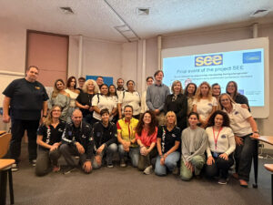 Group picture of meeting participants, some visibly part of different aid organisations by their uniforms/shirts, before a projection identifying the event as part of the EU co-financed SEE project.
